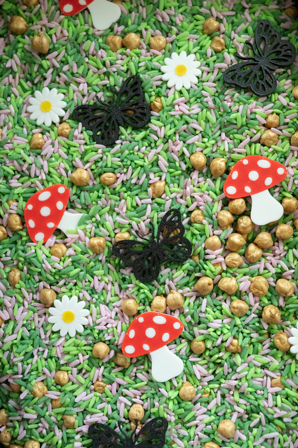 A close-up overhead view of a decorative Sensory bin filler mix featuring green and pink sprinkles, gold-topped peanuts, white daisy flowers, red-and-white mushroom shapes, and black butterfly decorations scattered across the surface.